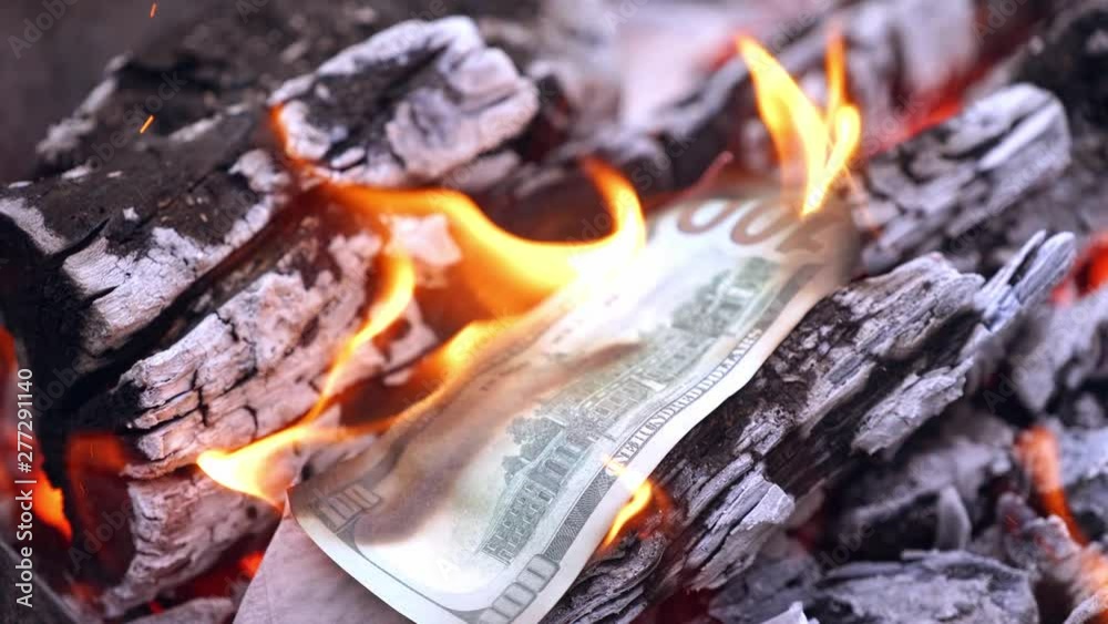 One hundred dollar bill burns in firewood. Someone's hand puts banknote of american money into fire on the background of burning wood. Close-up. One hundred dollar bill burns in firewood. Someone's hand puts banknote of american money into fire on the background of burning wood. Close-up.