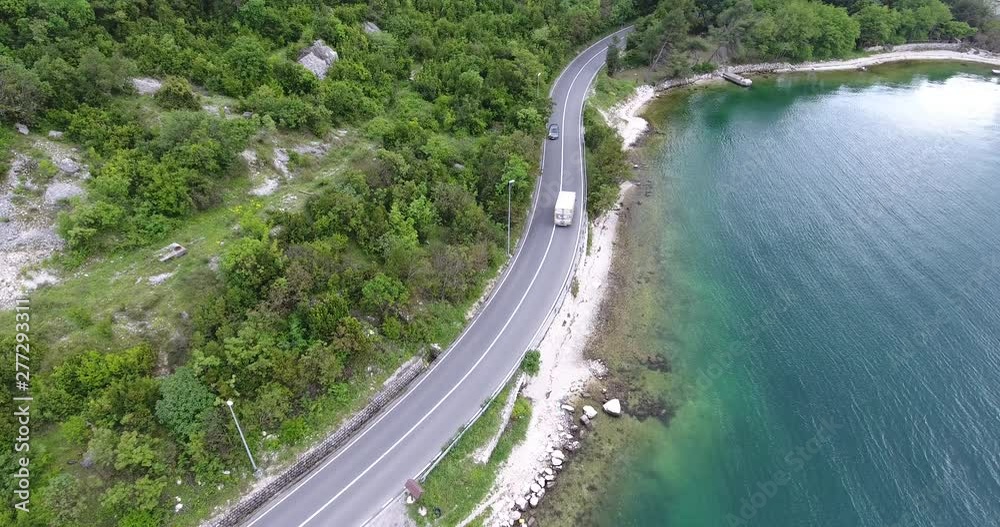 The quadcopter is flying over a narrow asphalt road running along the sea shore. We can see the turquoise sea and the seashore covered with green trees on the other side of the road The quadcopter is flying over a narrow asphalt road running along the sea shore. We can see the turquoise sea and the seashore covered with green trees on the other side of the road