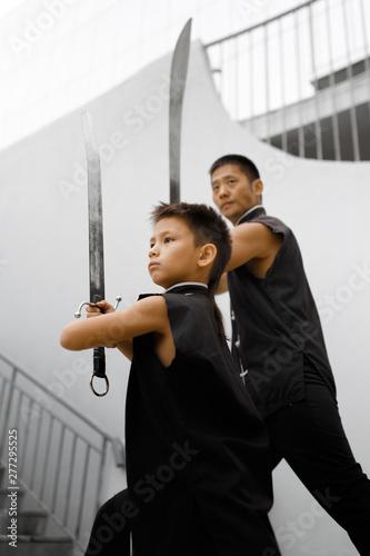 Father and son are engaged in Wushu in the city. The photo illustrates a healthy lifestyle and sport. The father trains the son. Close-up of son and father with swords.