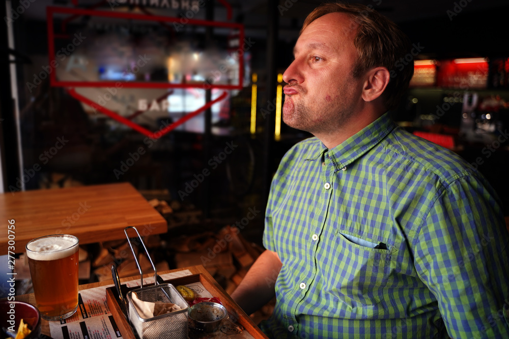 a man in a green shirt sitting at a bar table drinking beer and eating French fries with a disgruntled face a man in a green shirt sitting at a bar table drinking beer and eating French fries with a disgruntled face