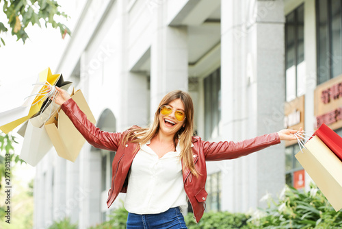 Portrait of happy excited young woman in sunglasses raising hands with paper-bags after shopping in city