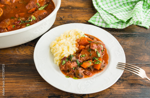 Coq au Vin, traditional French recipe of chicken braised in red wine with carrot and mushrooms. Served with mashed potatoes. White casserole and plate on wooden table