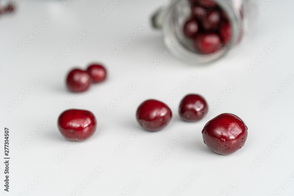 Red juicy cherries scattered from out the blurred glass jar on a white table