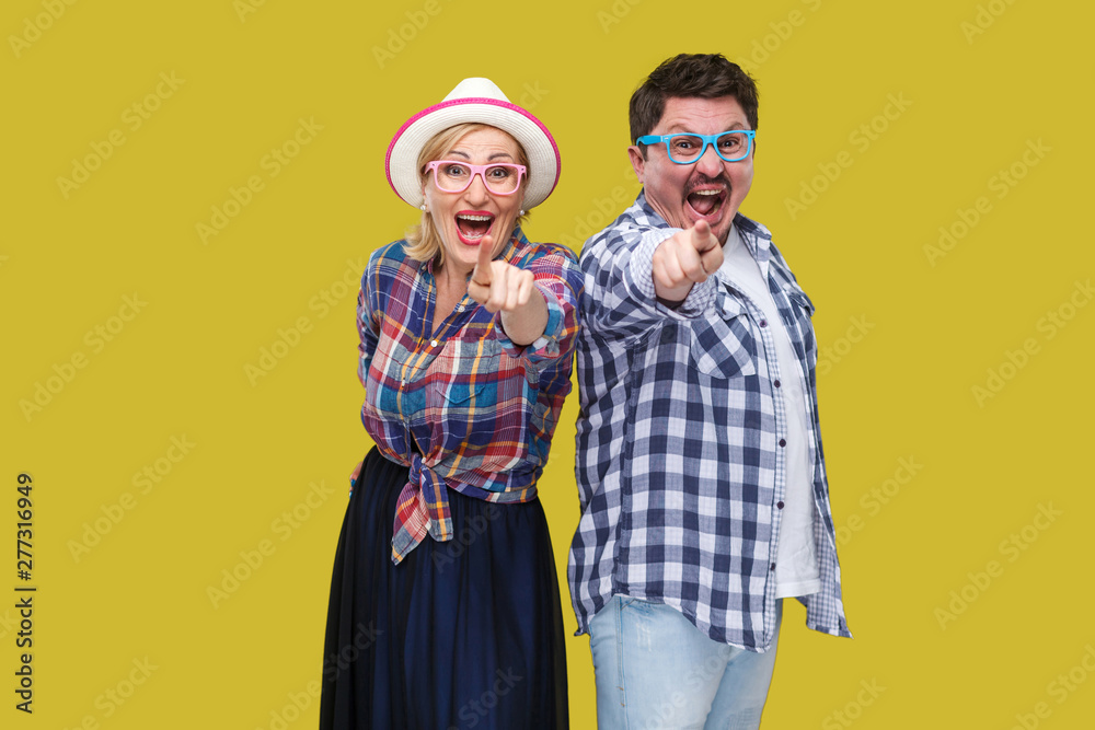 Couple of friends, adult man and woman in casual checkered shirt standing together back to back pointing finger, looking amazed, surprised or laughing. Indoor,isolated,studio shot, yellow background
