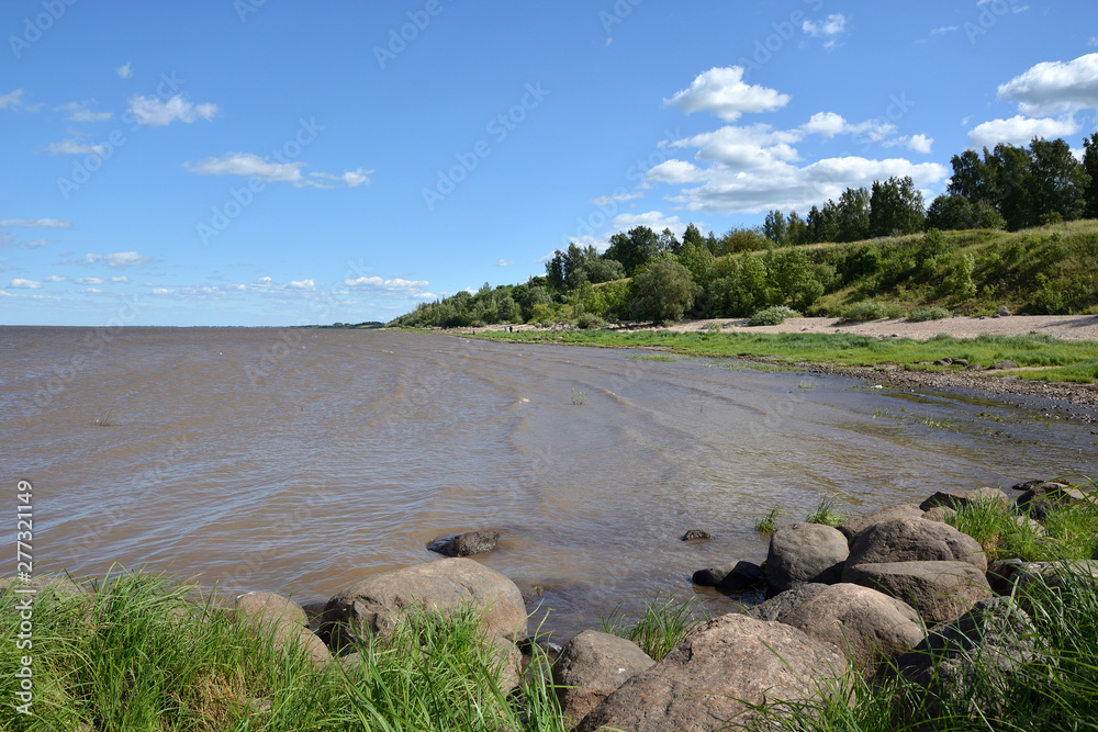 Summer landscape of a lake with stones in the foreground and a wooded and hilly shore in the background with a blue cloudy sky. Lake Ilmen Novgorod region