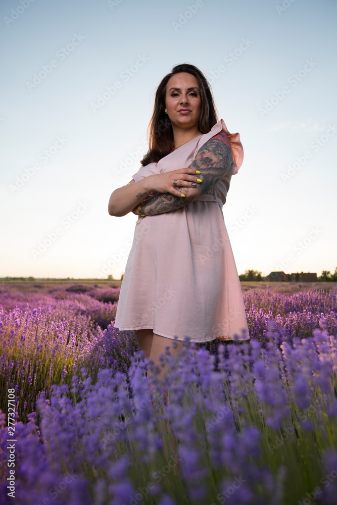 Young, beautiful, pretty girl with tattoos in a pastel dress. Woman standing amidst blooming colorful flowers in the lavender field against a bright blue sky during a warm summer sunset.