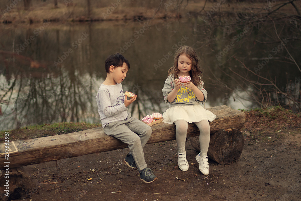 boy and girl eating doughnuts sitting on the wooden bench in the park