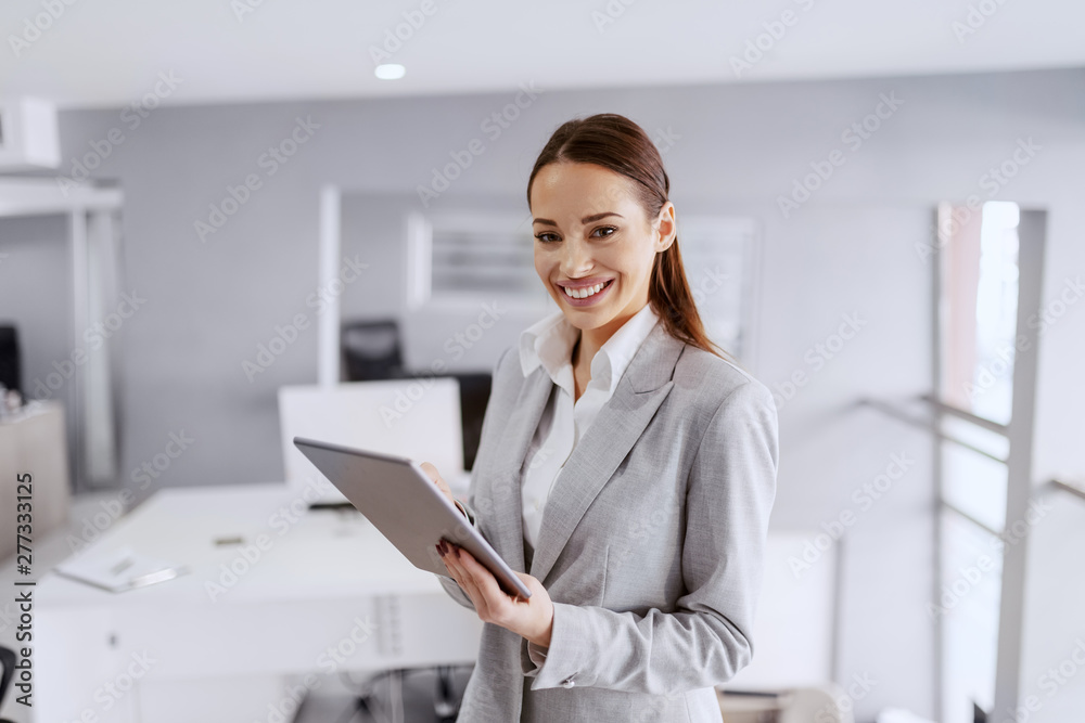 Gorgeous smiling Caucasian businesswoman in formal wear standing in office and using tablet. Gorgeous smiling Caucasian businesswoman in formal wear standing in office and using tablet.