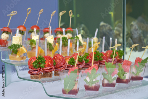 Different types of canapes in transparent glasses and wooden skewers on a glass step support on blue background and tank