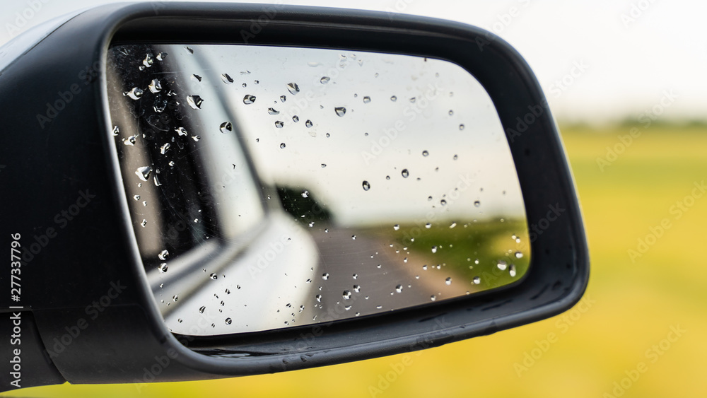 raindrops on side rear-view mirror on a car in a raining day. drops of rain on car window. sunset, sun light, green field over rainy wet road. driving in bad weather. raindrops on side rear-view mirror on a car in a raining day. drops of rain on car window. sunset, sun light, green field over rainy wet road. driving in bad weather.