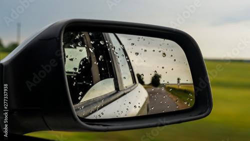 raindrops on side rear-view mirror on a car in a raining day. drops of rain on car window. sunset, sun light, green field over rainy wet road. driving in bad weather.