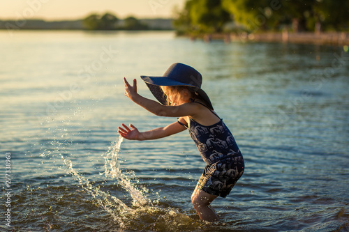 Fototapeta Naklejka Na Ścianę i Meble -  little girl in a hat by the river in summer splashes water