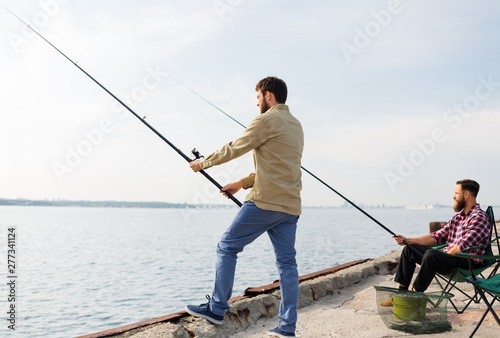 leisure and people concept - male friends with fishing rods on pier at sea