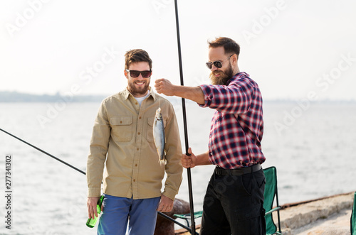 leisure and people concept - happy male friends with fishing rods, fish and beer talking on pier at sea