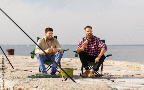 leisure and people concept - happy friends fishing and eating sandwiches on pier at sea