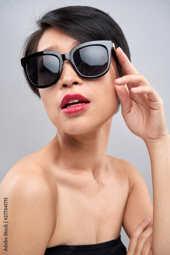 Portrait of Asian fashion girl with short black hair posing at camera in stylish sunglasses isolated on grey background