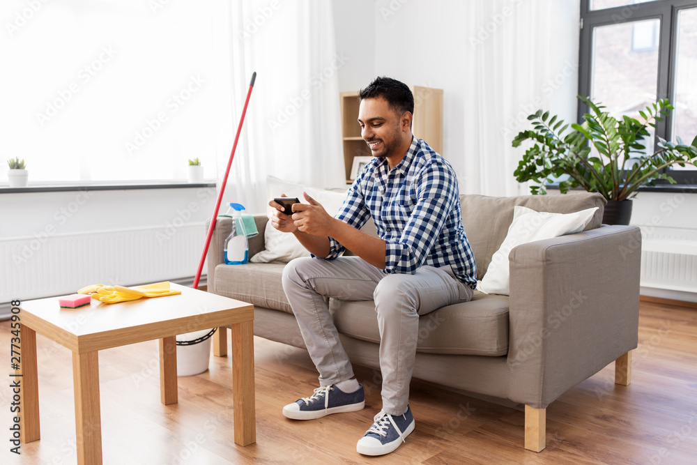 household and technology concept - smiling indian man playing game on smartphone after cleaning home
