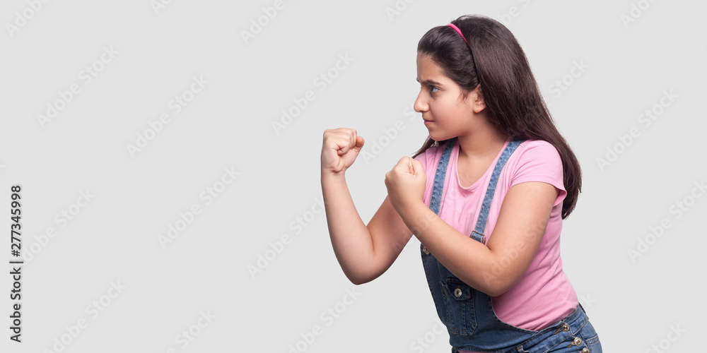 Profile side view portrait of serious or angry young girl in pink t-shirt and blue overalls standing with fists boxing hands and ready to attack. indoor studio shot, isolated on light gray background. Profile side view portrait of serious or angry young girl in pink t-shirt and blue overalls standing with fists boxing hands and ready to attack. indoor studio shot, isolated on light gray background.
