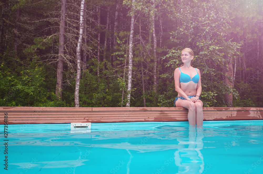 Caucasian girl in a blue swimsuit sits near the forest pool in the open. She looks away and smiles. The girl is on the right side of the frame. The concept of summer holidays, travel. Place for text.