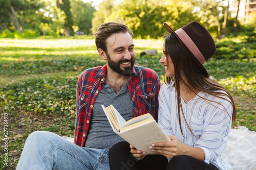 Image of joyful couple man and woman reading book while resting in green park