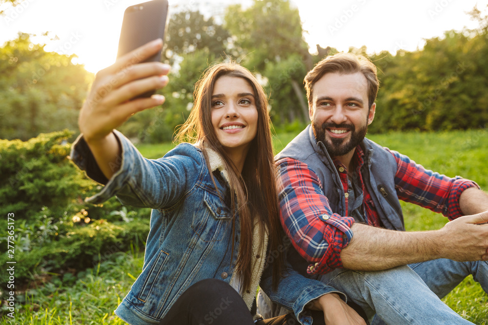 Image of cute couple man and woman taking selfie photo on cellphone while resting in green park