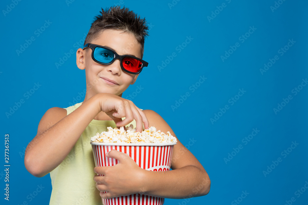the boy carefully watches a movie in 3D glasses, with a bucket of popcorn, the concept of cinema and entertainment, picks up popcorn, on a blue background