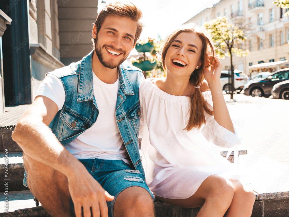 Obraz premium Smiling beautiful girl and her handsome boyfriend. Woman in casual summer dress and man in jeans clothes. Happy cheerful family. Sitting on stairs on the street background.Hugging couple