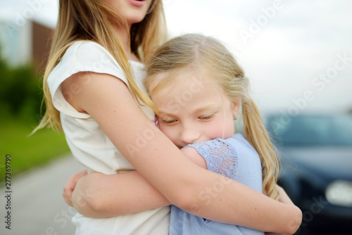 Two adorable sisters laughing and hugging on warm and sunny summer day in a city.