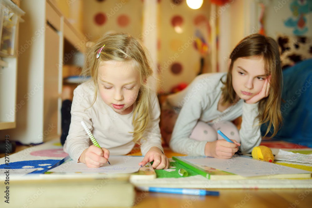 Two cute little sisters writing letters together at home. Older sister helping youngster with her homework. Two cute little sisters writing letters together at home. Older sister helping youngster with her homework.
