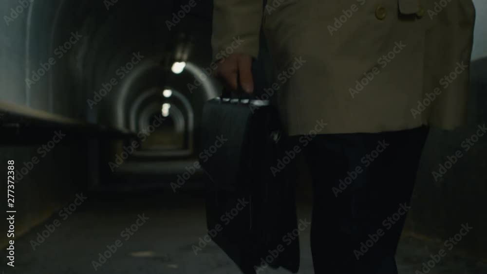 Low-section front view shot of man in a coat with case walking through the tunnel in the evening Low-section front view shot of man in a coat with case walking through the tunnel in the evening
