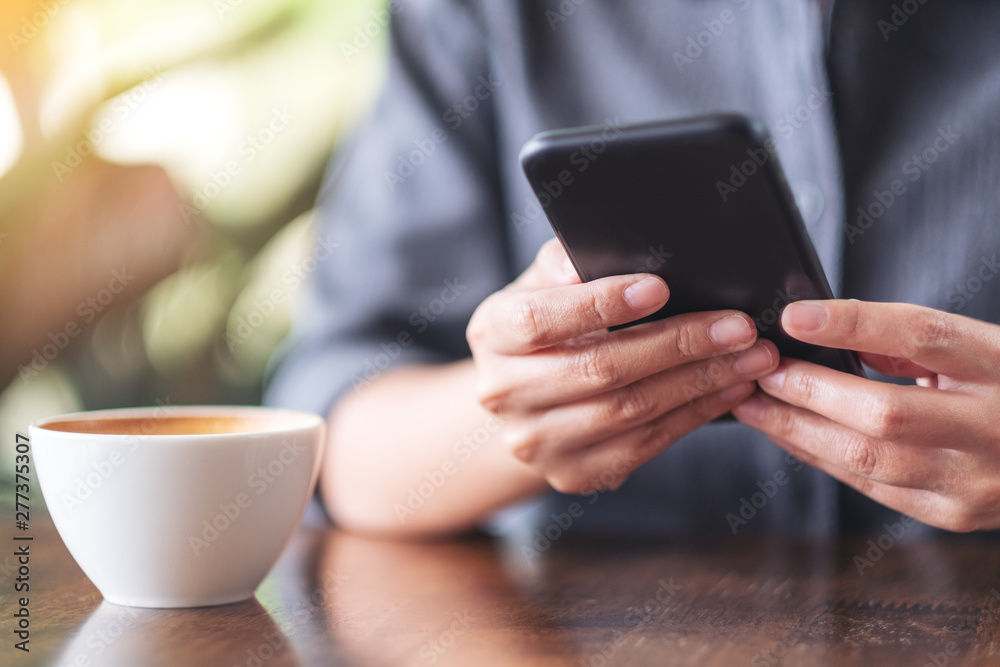 Closeup image of a woman holding and using mobile phone with coffee cup on wooden table in cafe Closeup image of a woman holding and using mobile phone with coffee cup on wooden table in cafe