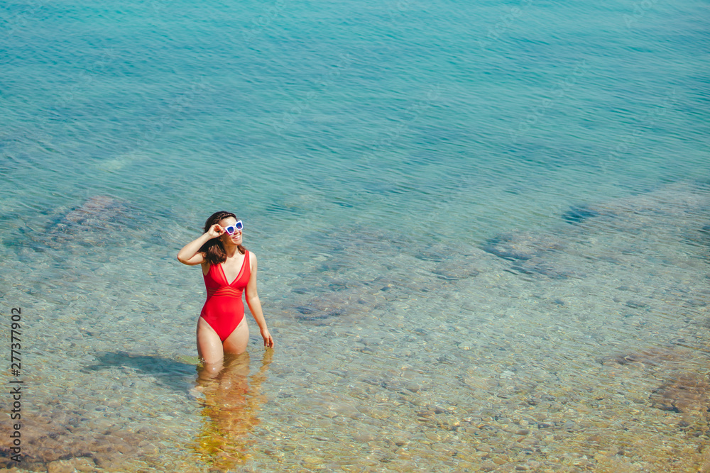 woman in red swimsuit in white sunglasses standing in sea. copy space. summer vacation. yacht on background