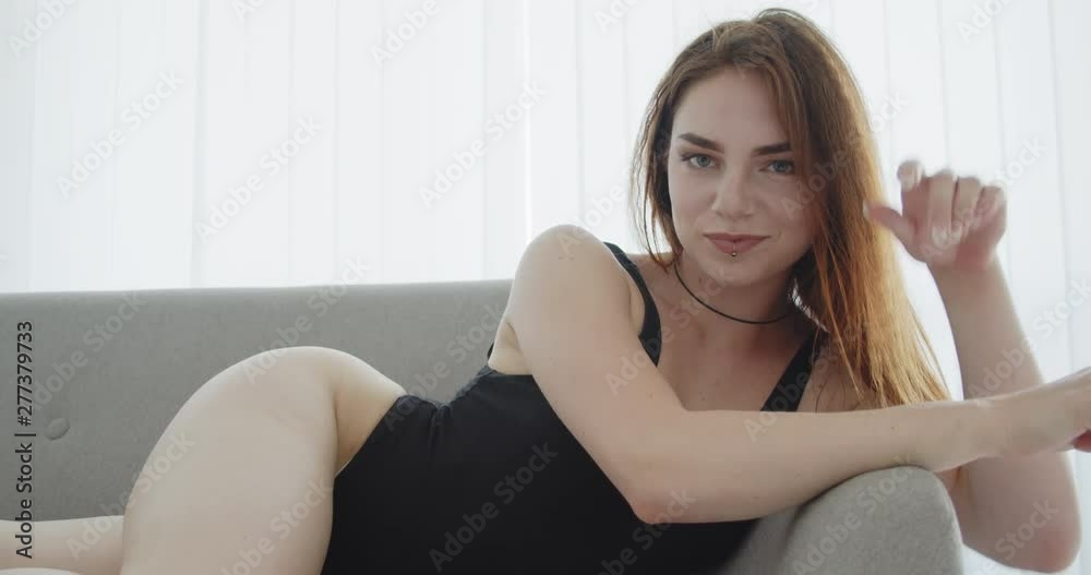 Closeup shoot of young sexy caucasian female in black bodysuit laying on the couch smiling and posing looking at camera indoors Closeup shoot of young sexy caucasian female in black bodysuit laying on the couch smiling and posing looking at camera indoors