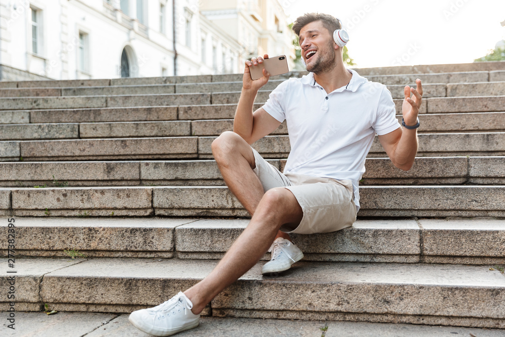 Portrait of happy young man wearing headphones holding smartphone and singing outdoors Portrait of happy young man wearing headphones holding smartphone and singing outdoors