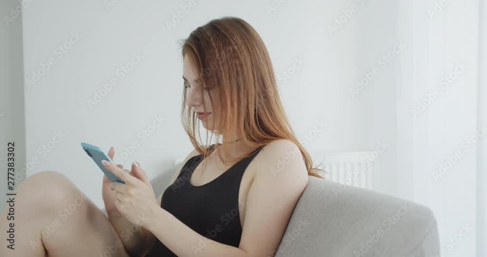 Closeup shoot of young sexy caucasian female in black bodysuit using the phone turning to camera and posing cheerfully sitting on the couch indoors Closeup shoot of young sexy caucasian female in black bodysuit using the phone turning to camera and posing cheerfully sitting on the couch indoors