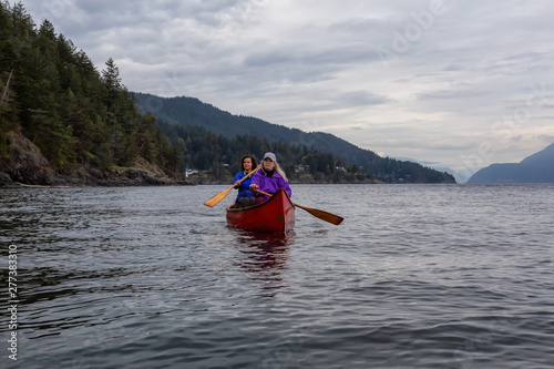 Couple adventurous female friends on a red canoe are paddling in the Howe Sound during a cloudy sunset. Taken near Bowen Island, West of Vancouver, BC, Canada.