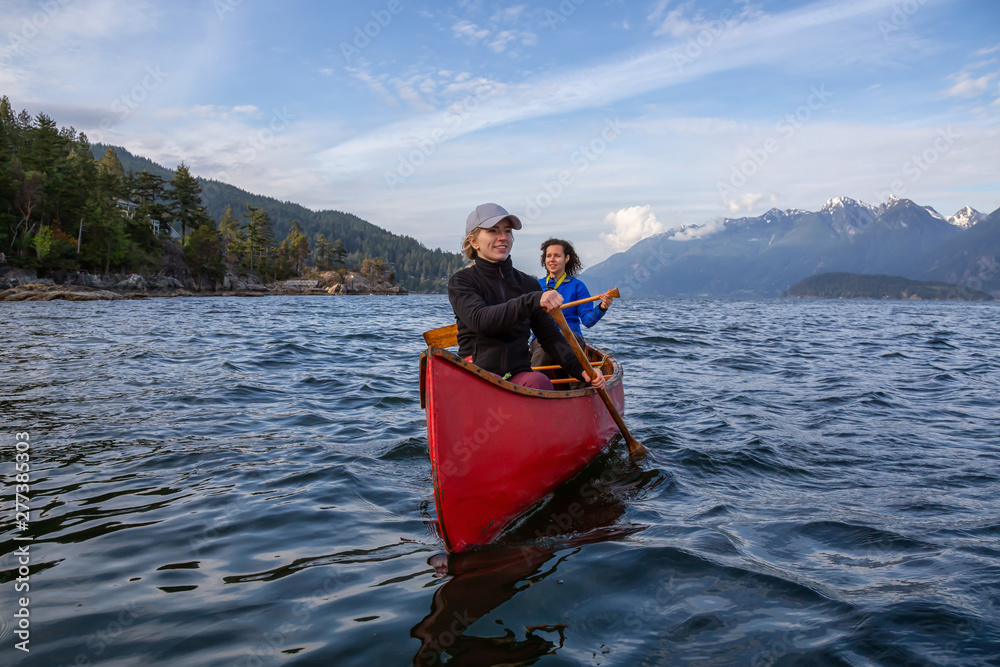 Couple adventurous female friends on a red canoe are paddling in the Howe Sound during a cloudy and sunny evening. Taken near Bowen Island, West of Vancouver, BC, Canada.