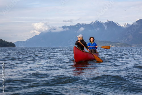 Couple adventurous female friends on a red canoe are paddling in the Howe Sound during a cloudy and sunny evening. Taken near Bowen Island, West of Vancouver, BC, Canada.
