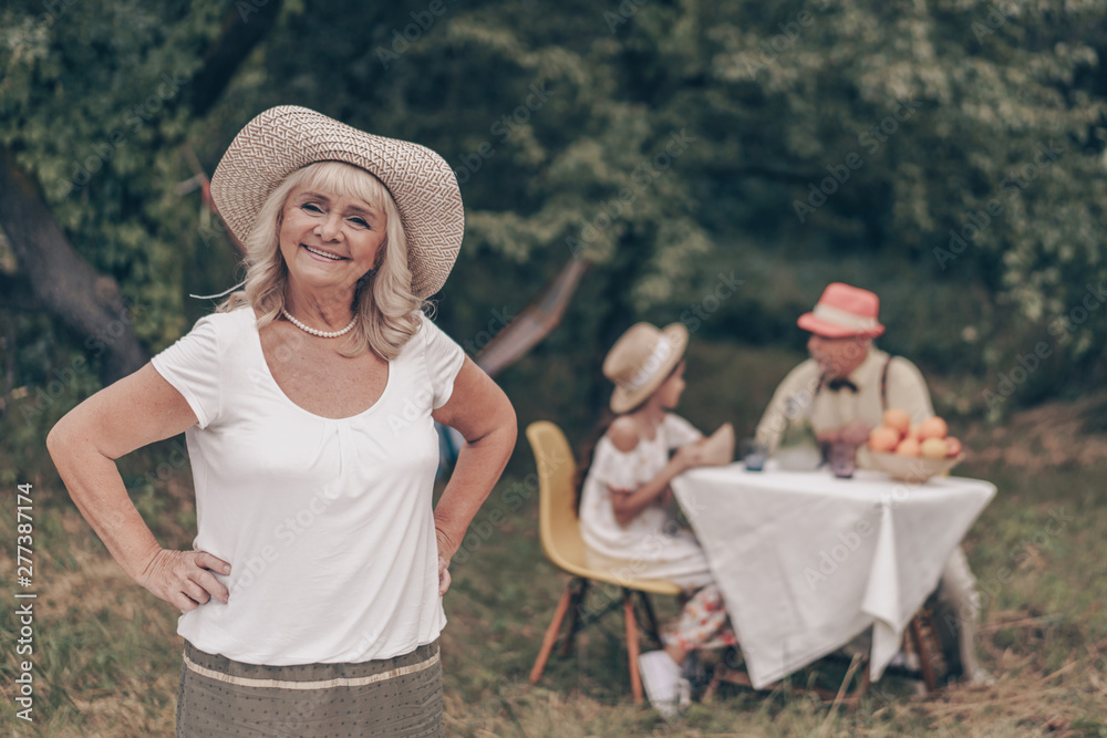 A beautiful portrait of a smiling grandmother in a T-shirt, skirt and hat in the garden. The happy granddaughter came to visit her grandfather and sit at the table.