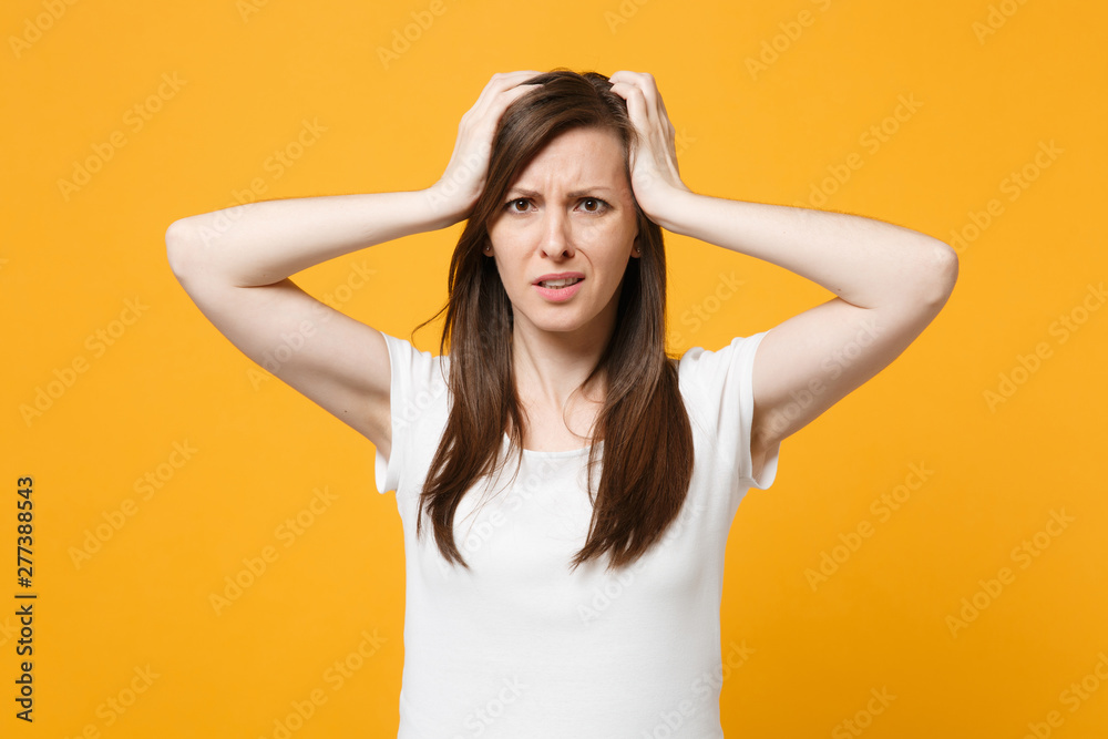 Portrait of preoccupied young woman in white casual clothes looking camera, putting hands on head isolated on yellow orange wall background in studio. People lifestyle concept. Mock up copy space.