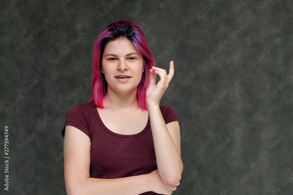 Portrait to the waist of a young pretty girl teenager in a burgundy T-shirt with beautiful purple hair on a gray background in the studio. Talking, smiling, showing hands with emotions. Portrait to the waist of a young pretty girl teenager in a burgundy T-shirt with beautiful purple hair on a gray background in the studio. Talking, smiling, showing hands with emotions.