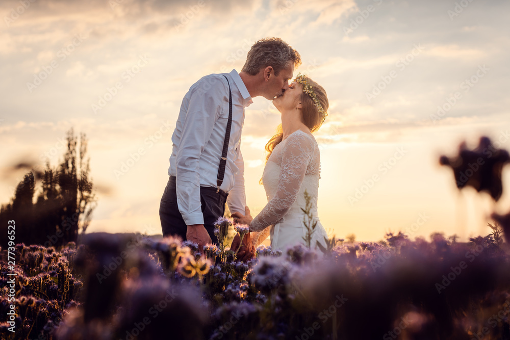 Hochzeitspaar küsst sich zum Sonnenuntergang auf der Wiese nach der Hochzeit  - obrazy, fototapety, plakaty 