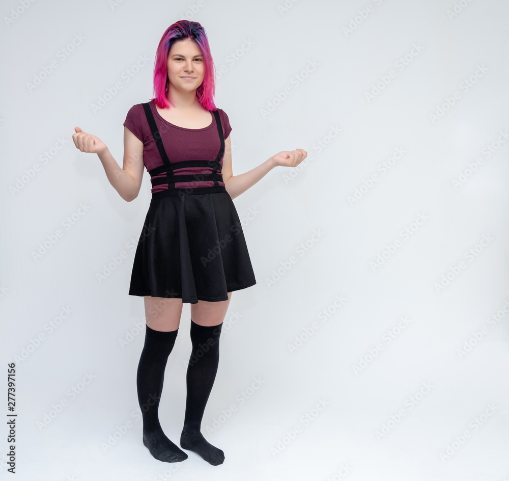 Full-length portrait of young pretty teenager girl in burgundy T-shirt and black dress with beautiful purple hair on a white background in the studio. Talking, smiling, showing hands with emotions.