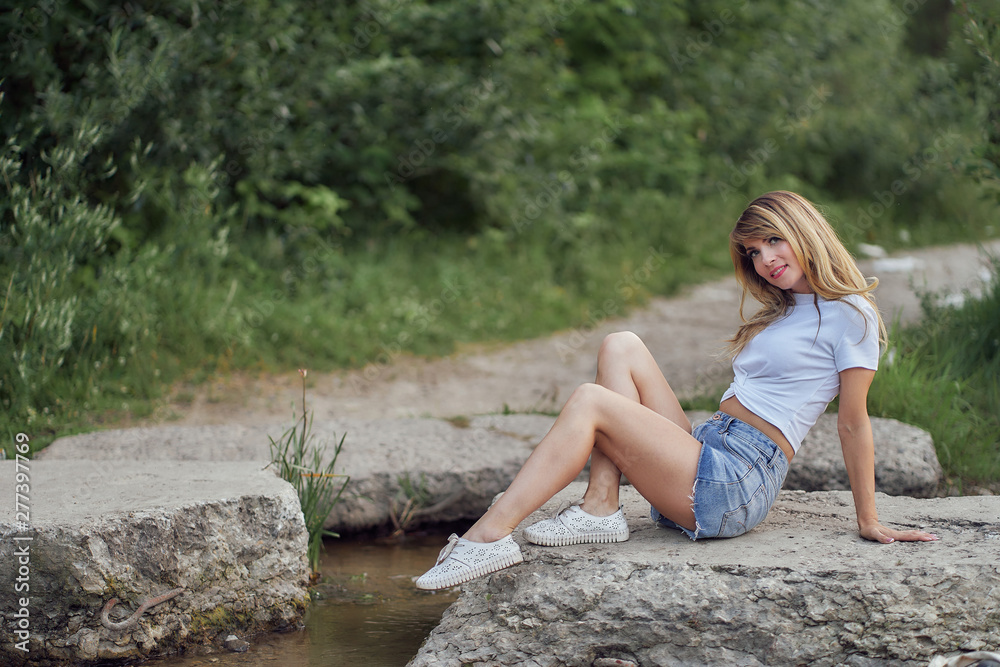 slim girl in shorts and white T-shirt sits on the rocks near the water on the lake slim girl in shorts and white T-shirt sits on the rocks near the water on the lake