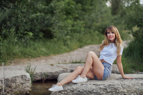 slim girl in shorts and white T-shirt sits on the rocks near the water on the lake