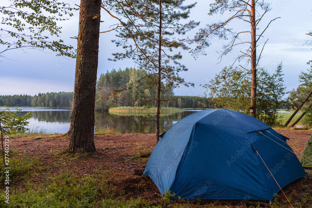 Camping tourism and a blue tent near a picturesque lake outdoor. Summer evening sunset sky. Camping and travel concept.