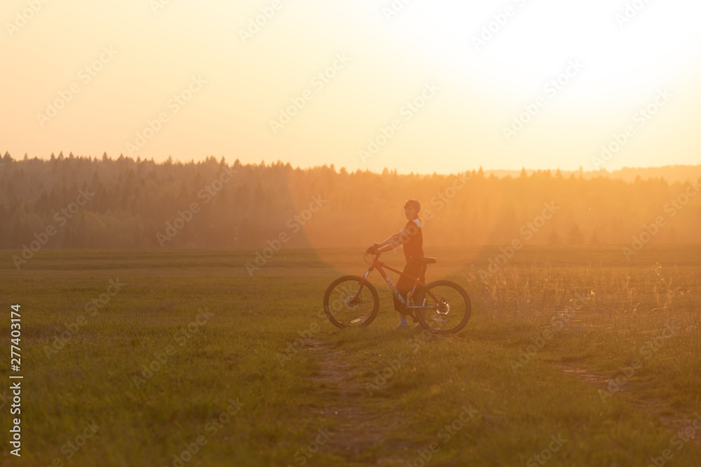 Obraz premium A girl rides a bicycle on the road against the backdrop of a bright sunset and yellow sun
