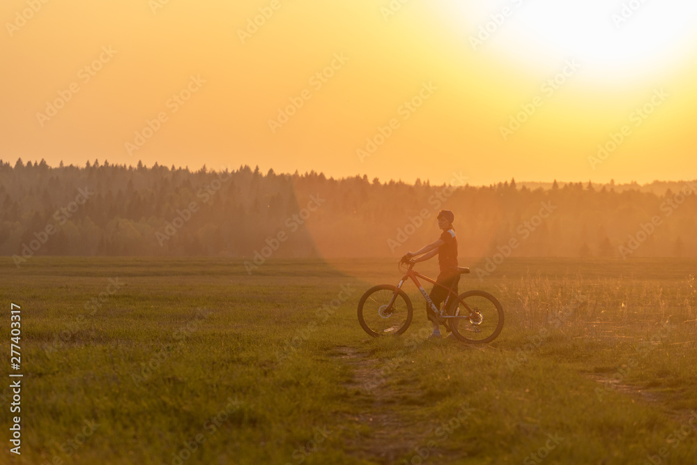 Obraz premium A girl rides a bicycle on the road against the backdrop of a bright sunset and yellow sun