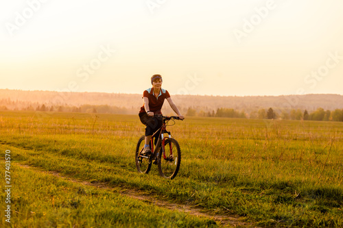 Fototapeta Naklejka Na Ścianę i Meble -  A girl rides a bicycle on the road against the backdrop of a bright sunset and yellow sun