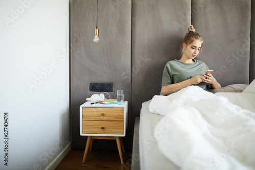 Young relaxed woman sitting in bed under banket while scrolling in smartphone © Seventyfour